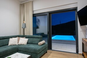 A modern living room with a green sofa, a marble coffee table, a TV, and a sliding glass door leading to an outdoor swimming pool area. - Relaxed Family Villa in Naklice (Naklice)