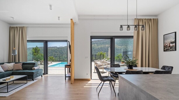Una sala de estar moderna con sofá, mesa de centro y comedor. Hay un ventanal con vista a una piscina y colinas.