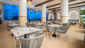 A spacious restaurant with high ceilings, large windows, and a view of an aquarium. Tables are set with chairs, and the floor is tiled. - Beachfront 1bedroom Apartment (Choeng Thale)