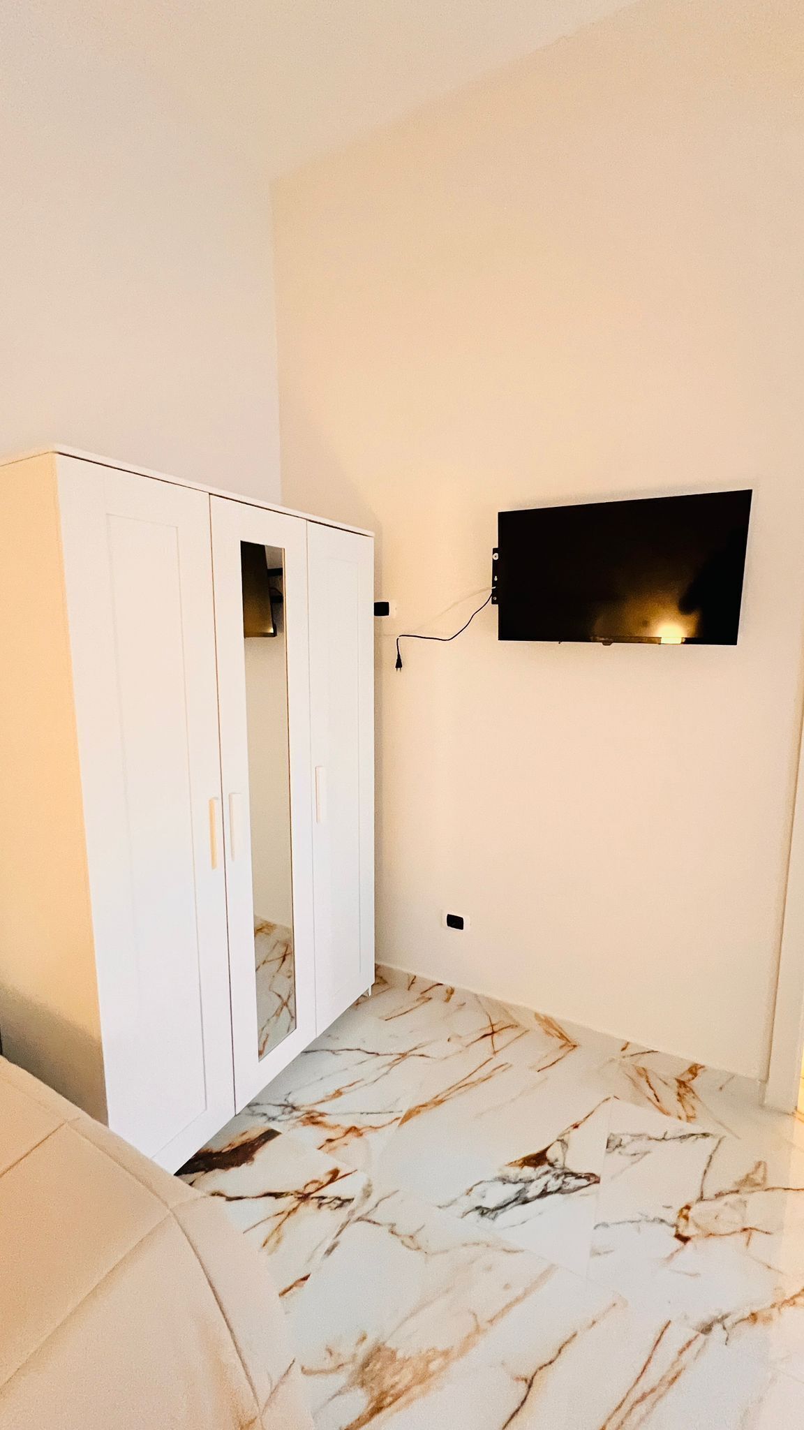 Ein Zimmer mit weißem Kleiderschrank, einem flachbildfernseher an der Wand und einem Boden mit Marmoroptik.