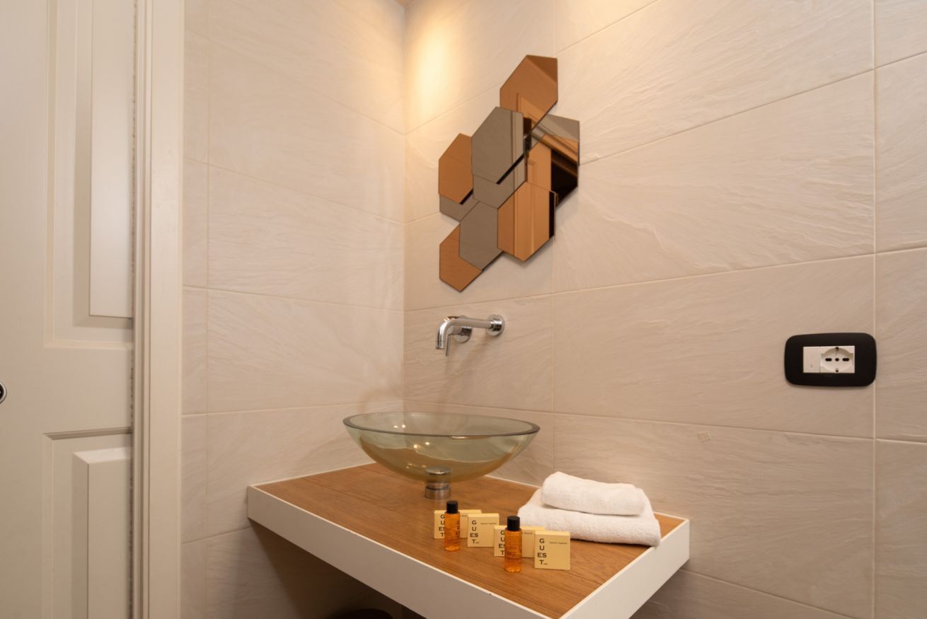 Un baño moderno con un lavabo de vidrio, un mueble de tocador de madera y un espejo geométrico en la pared.