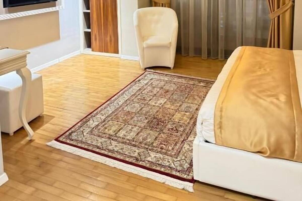 Habitación de hotel con cama, un sillón blanco, televisor montado en la pared y un ventanal con cortinas.