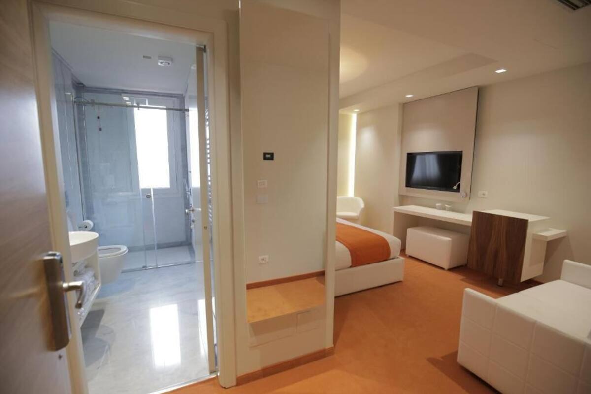 Habitación de hotel moderna con cabina de ducha de vidrio, sofá blanco, televisor montado en la pared y aparador de madera.