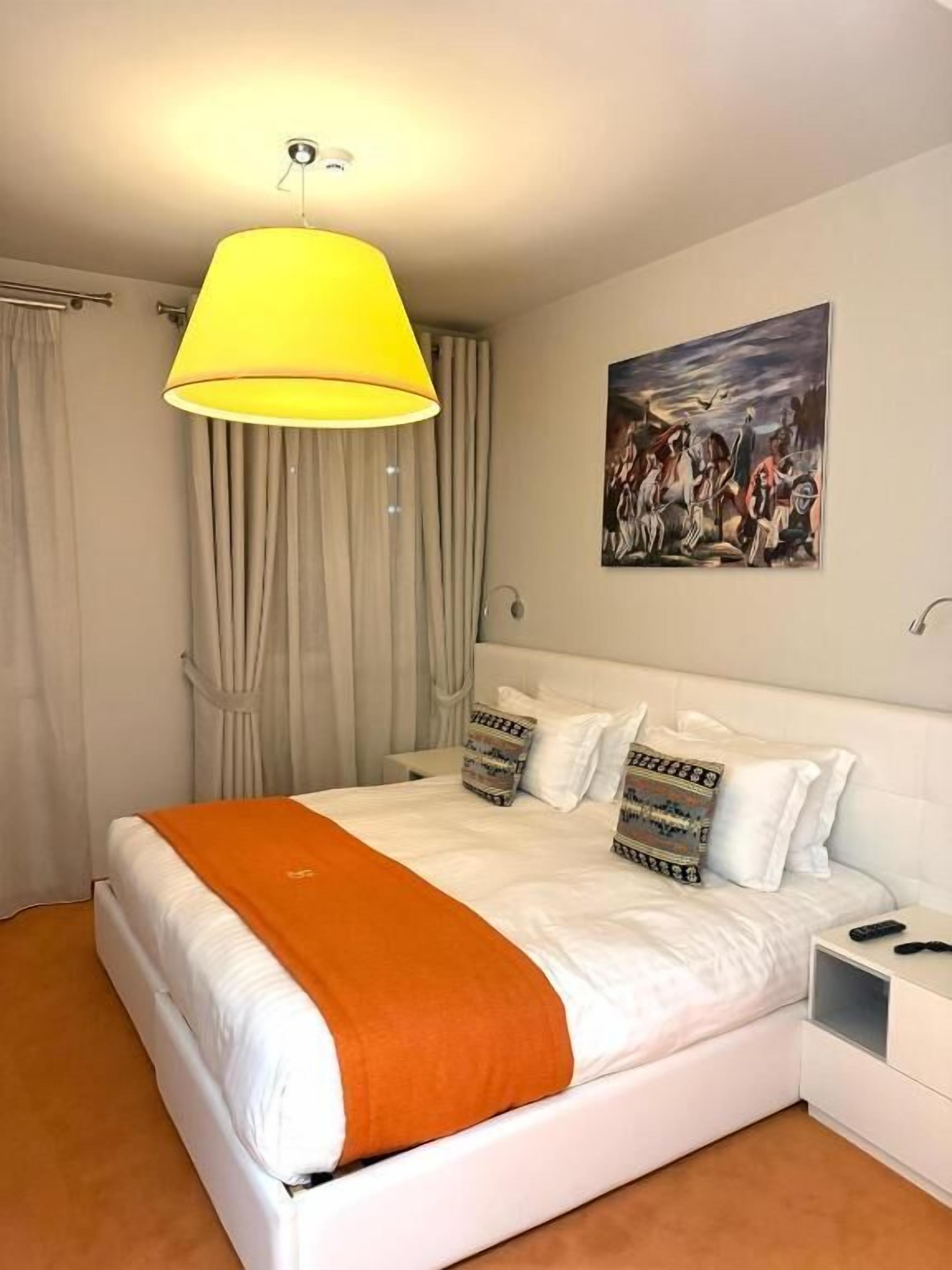 A hotel room with a bed, a nightstand, a lamp, curtains, and a painting on the wall.