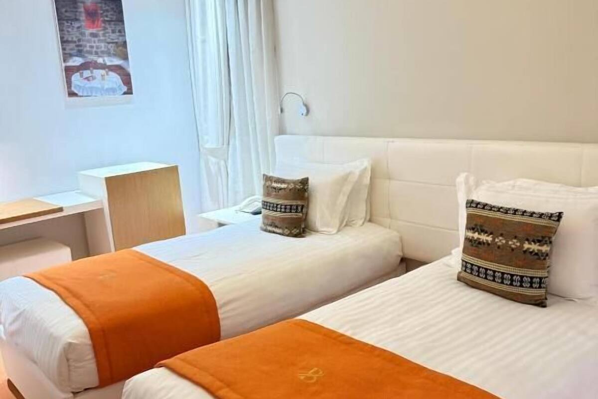 Habitación de hotel con dos camas, ropa de cama blanca, almohadones naranja, cabecera blanca, lámpara amarilla, cortina blanca y una pequeña mesa con una lámpara.