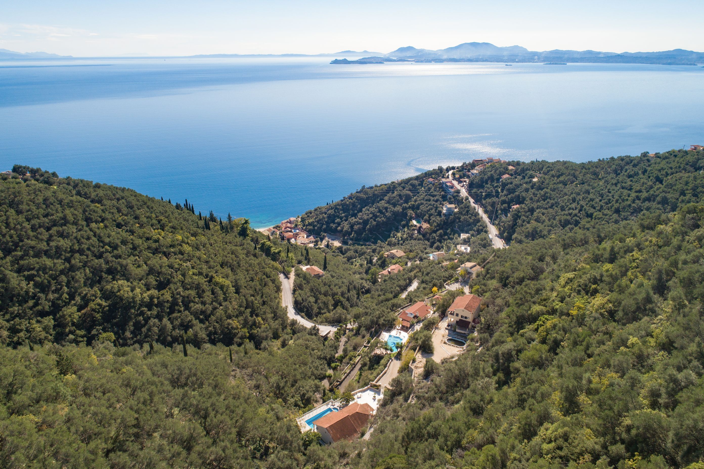 A coastal village with houses surrounded by dense forest, a clear view of the sea, and a winding road leading to the beach.