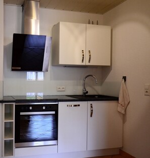 Een kleine keuken met witte kasten, een roestvrijstalen afzuigkap, een inbouwoven, een spoelbak met een kraan, en een handdoek aan de muur.