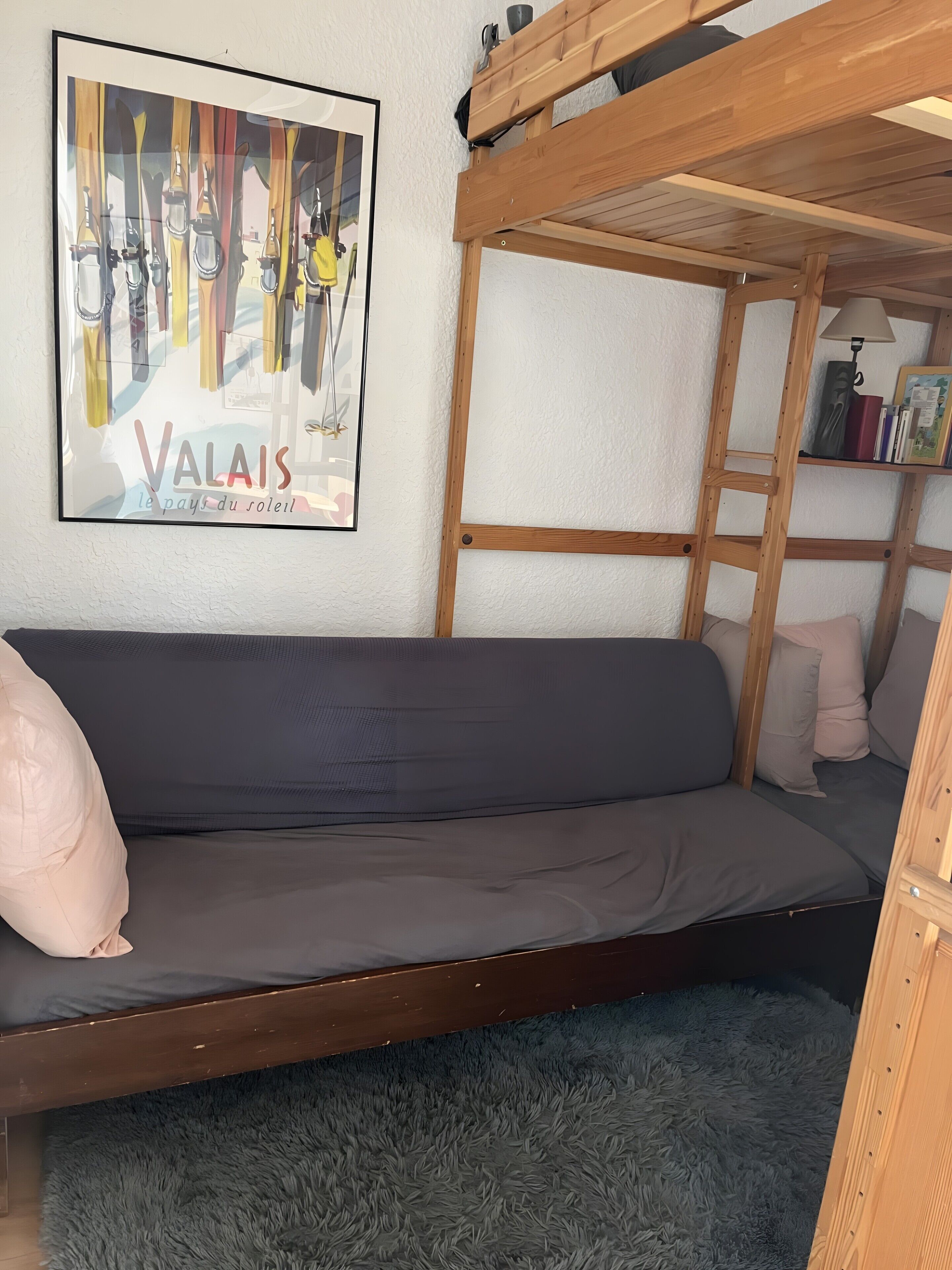 Ein Etagenbett mit grauem Lattenrost, ein gerahmtes Kunstwerk an der Wand und ein Regal mit Büchern und einer Lampe.