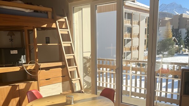 Una habitación con una mesa de comedor de madera, sillas rojas, una escalera de madera y ventanas amplias con vistas a edificios y montañas.