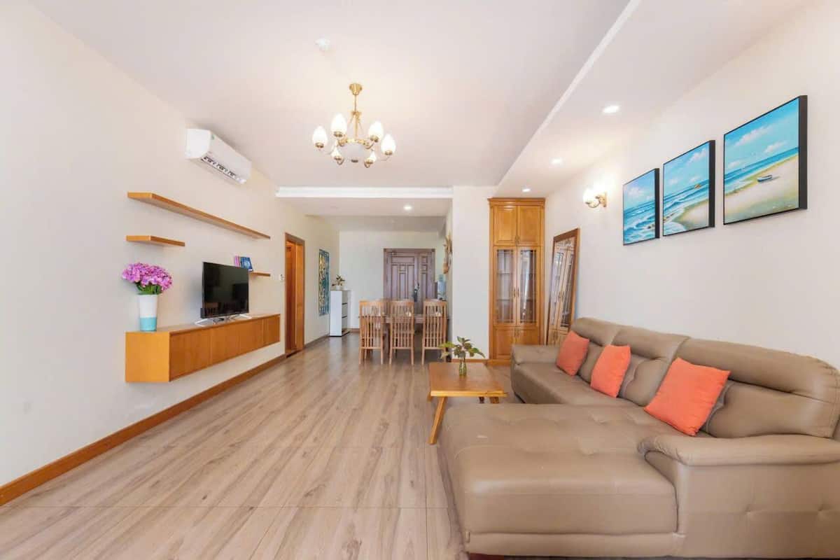 Un salon moderne avec un canapé beige, des étagères en bois, un téléviseur et, au fond, un coin repas.