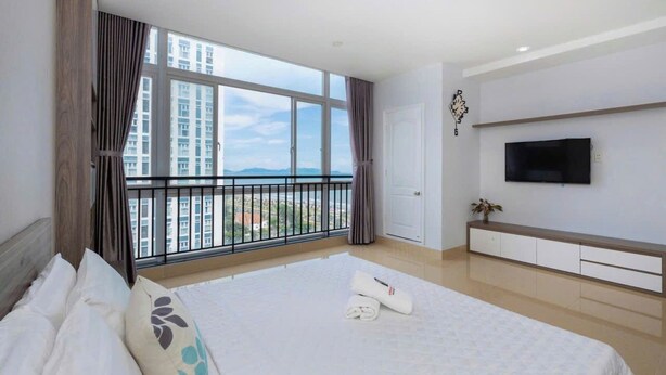 A hotel room with a large bed, a TV mounted on the wall, and a balcony with a view of buildings and mountains. - Oasky Vung Tau - AnNam Village (Ho Chi Minh City)