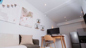 Un salon moderne avec un canapé, une table basse sur laquelle se trouve un saladier rempli de fruits, un téléviseur et, au fond, un coin cuisine.