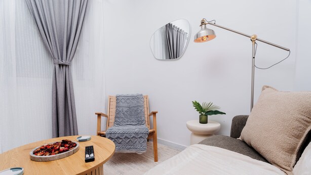 Une chambre à coucher bien rangée, avec un lit, une chaise, une table ronde sur laquelle se trouve un saladier rempli de fruits, une lampe et un miroir accroché au mur.