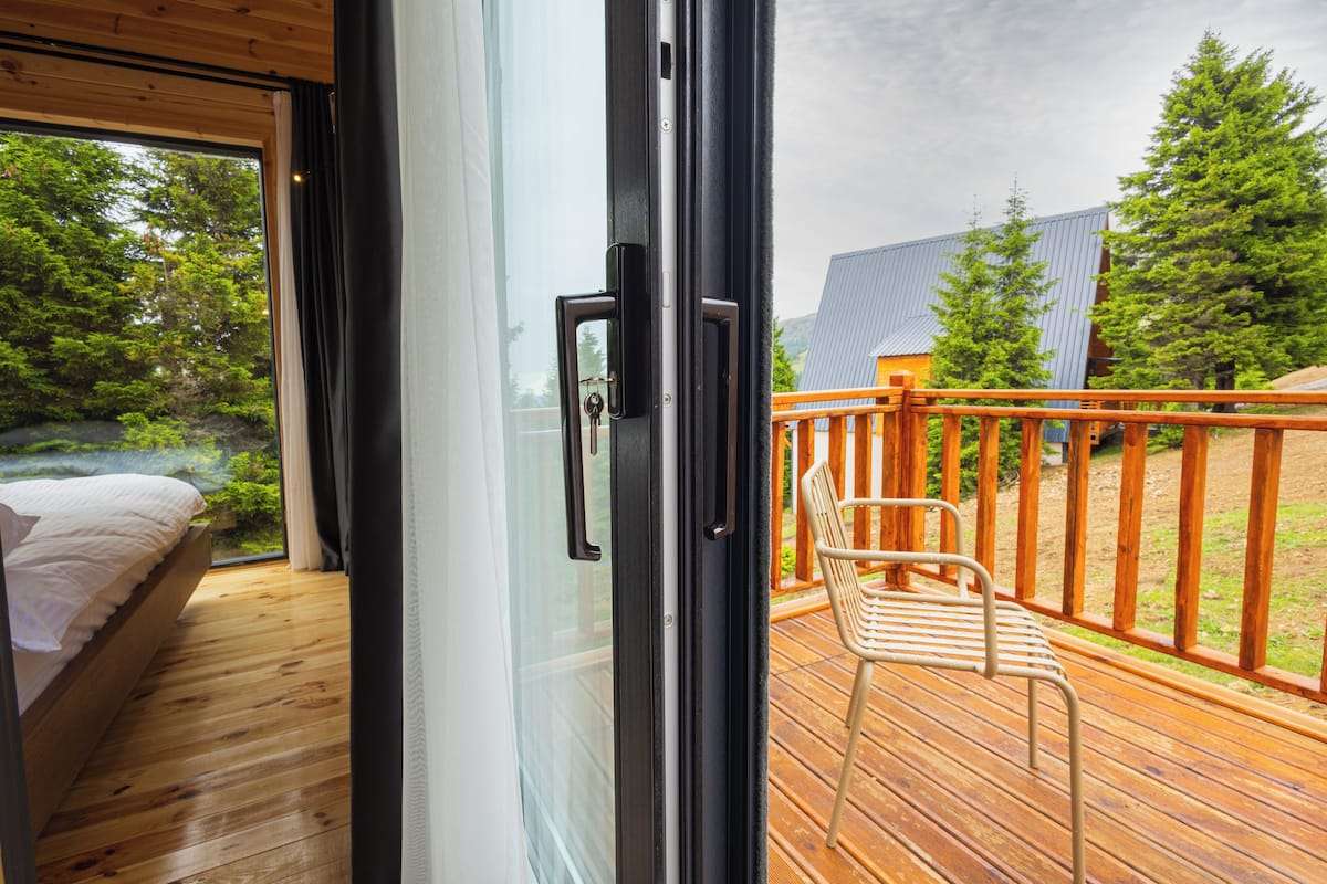 A bedroom with a wooden floor, a bed with white bedding, and a sliding glass door leading to an outdoor deck with a wooden railing and a chair.