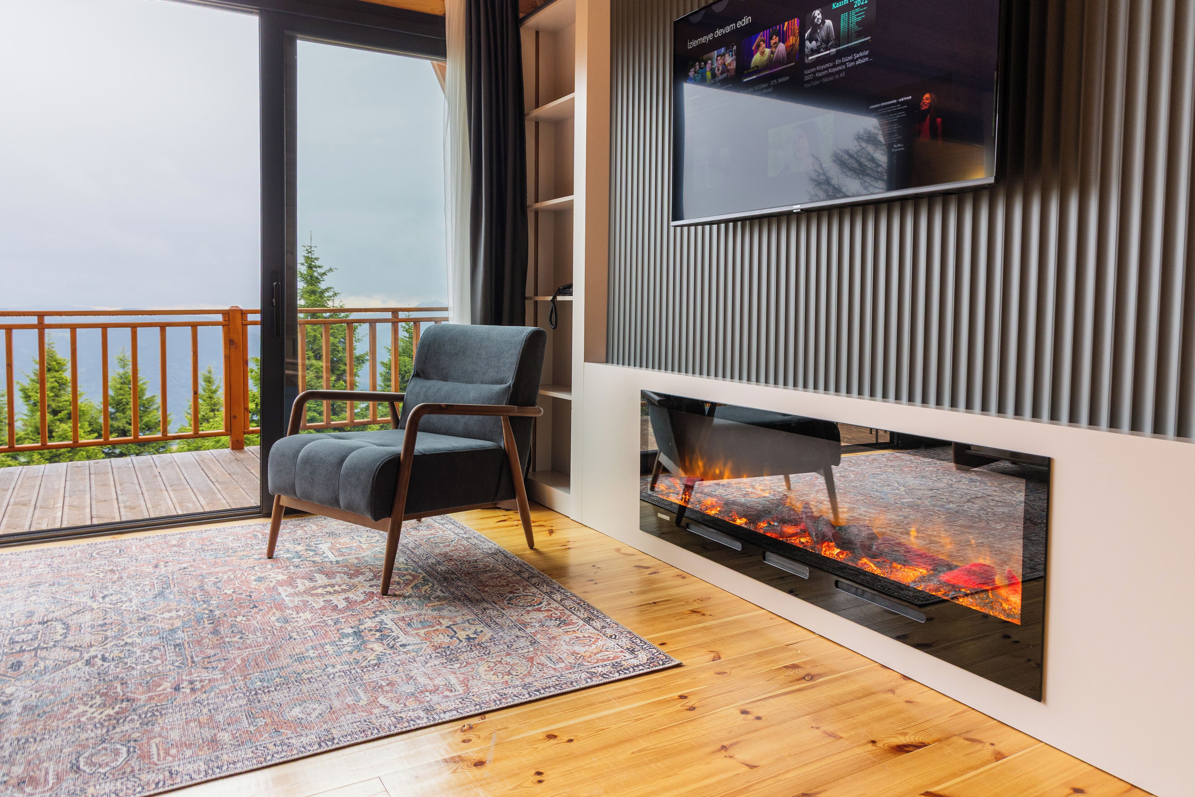 A modern living room with a TV mounted on the wall, a fireplace, a wooden deck, and a patterned rug.