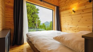 A bedroom with a wooden floor, a bed with white bedding, a nightstand, and a window with curtains. - TİMİRA DAĞ EVLERİ (Dereli)