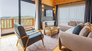 A spacious living room with a large window, a sofa, a coffee table, and a TV mounted on the wall. - TİMİRA DAĞ EVLERİ (Dereli)