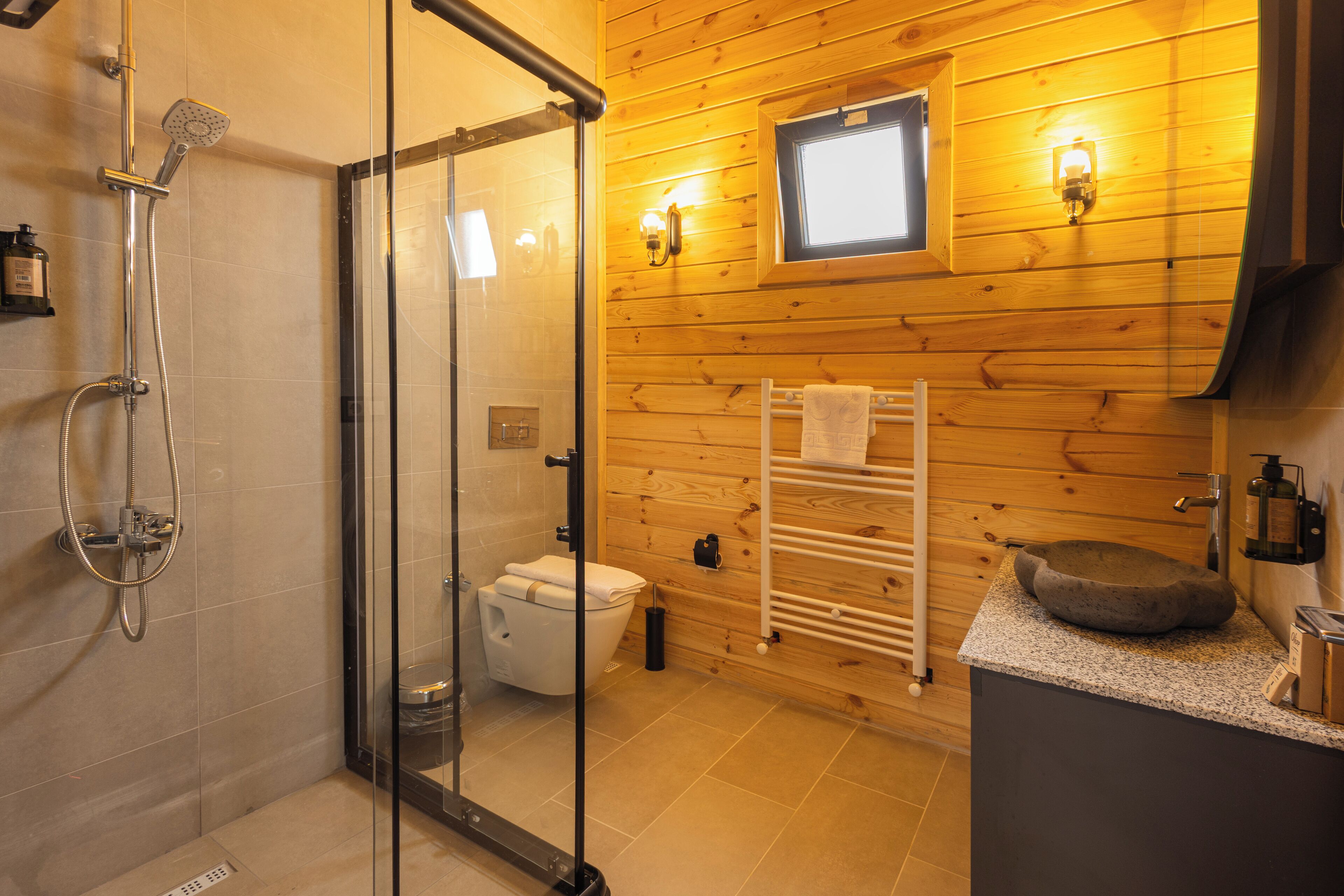 A modern bathroom with a glass shower enclosure, a wooden wall, a small window, and a stone sink.