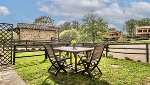 Eine Terrasse mit einem runden Tisch und Stühlen, ein kleines Steinhaus und ein von Bäumen gesäumtes, eingezäuntes Gelände.