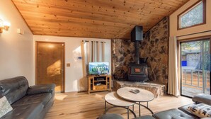 A cozy living room with a stone fireplace, wooden ceiling, and a television on a wooden stand. - Oglala Little Lodge - Cabin Getaway (South Lake Tahoe)