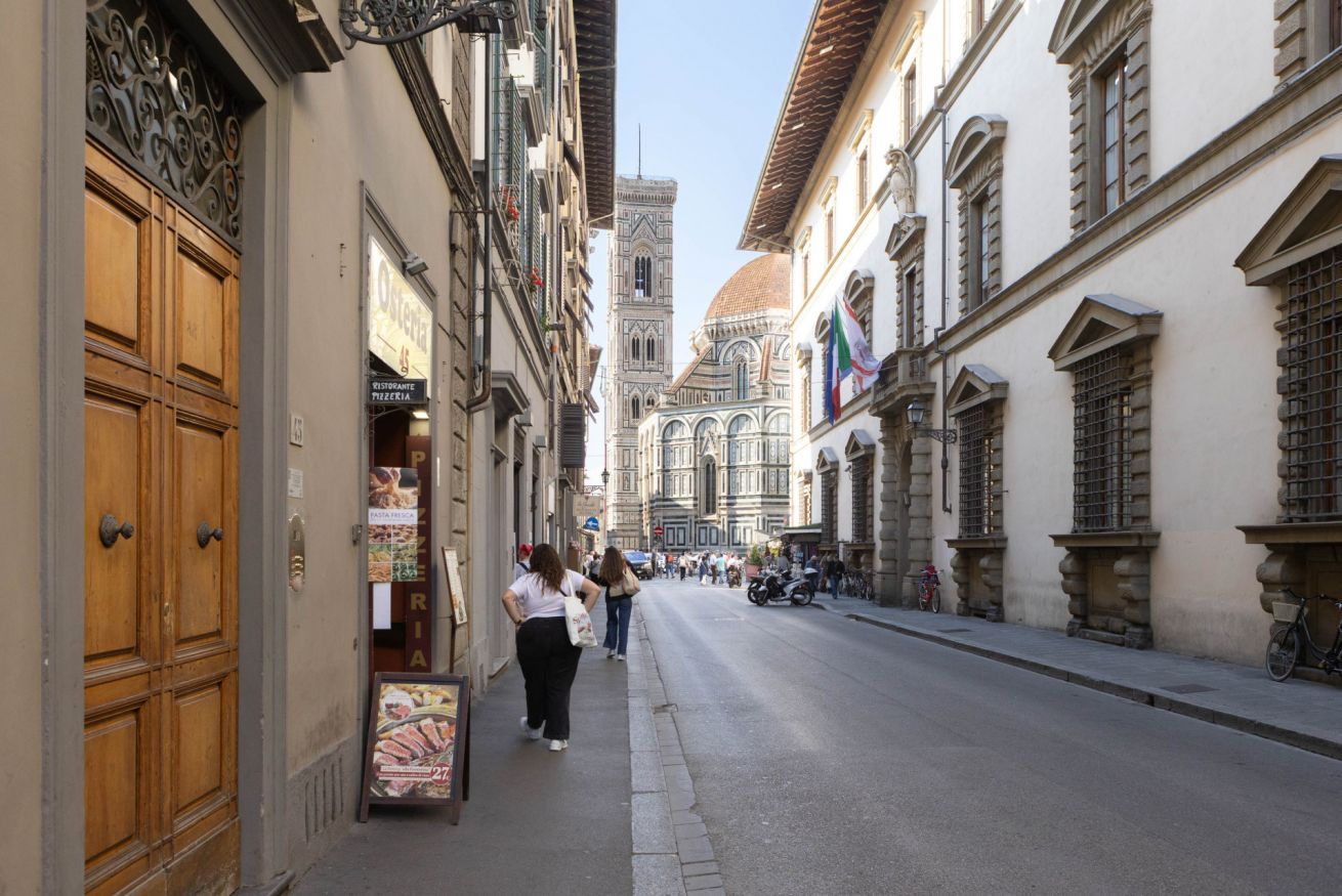 A narrow street lined with historic buildings, a prominent church dome in the background, and pedestrians walking along the sidewalk.