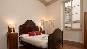 A bedroom with a wooden bed, two bedside tables with lamps, a window with shutters, and framed pictures on the wall. - Frescoed Duomo Retreat (Firenze)