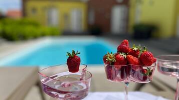 Un cadre agréable au bord d’une piscine, avec une vue dégagée sur celle-ci, un bâtiment jaune et une table sur laquelle reposent deux verres contenant une boisson rosée, chacun agrémenté d’une fraise.