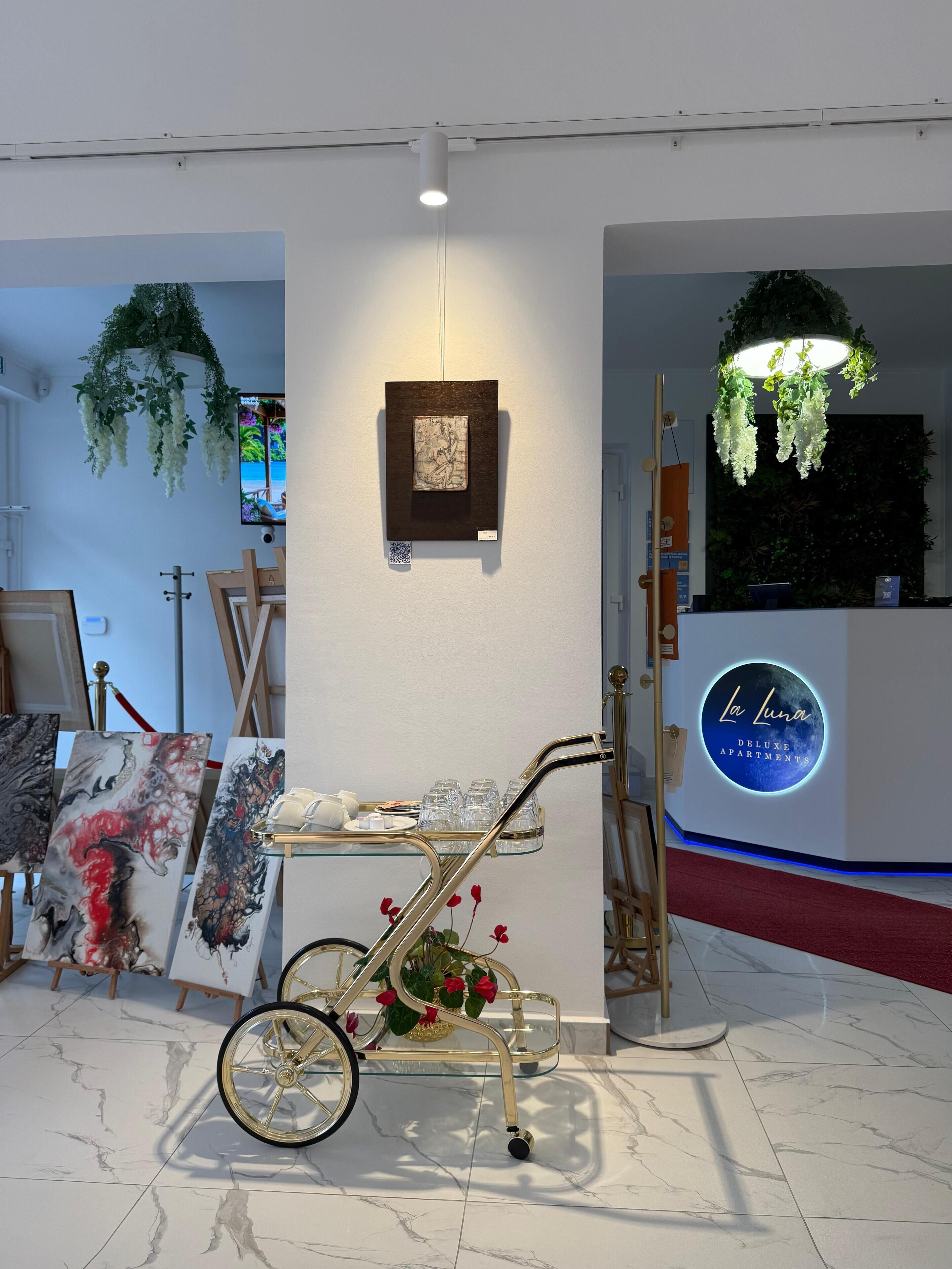 An art gallery with paintings on easels, a cart with glassware, and a reception desk with a sign.