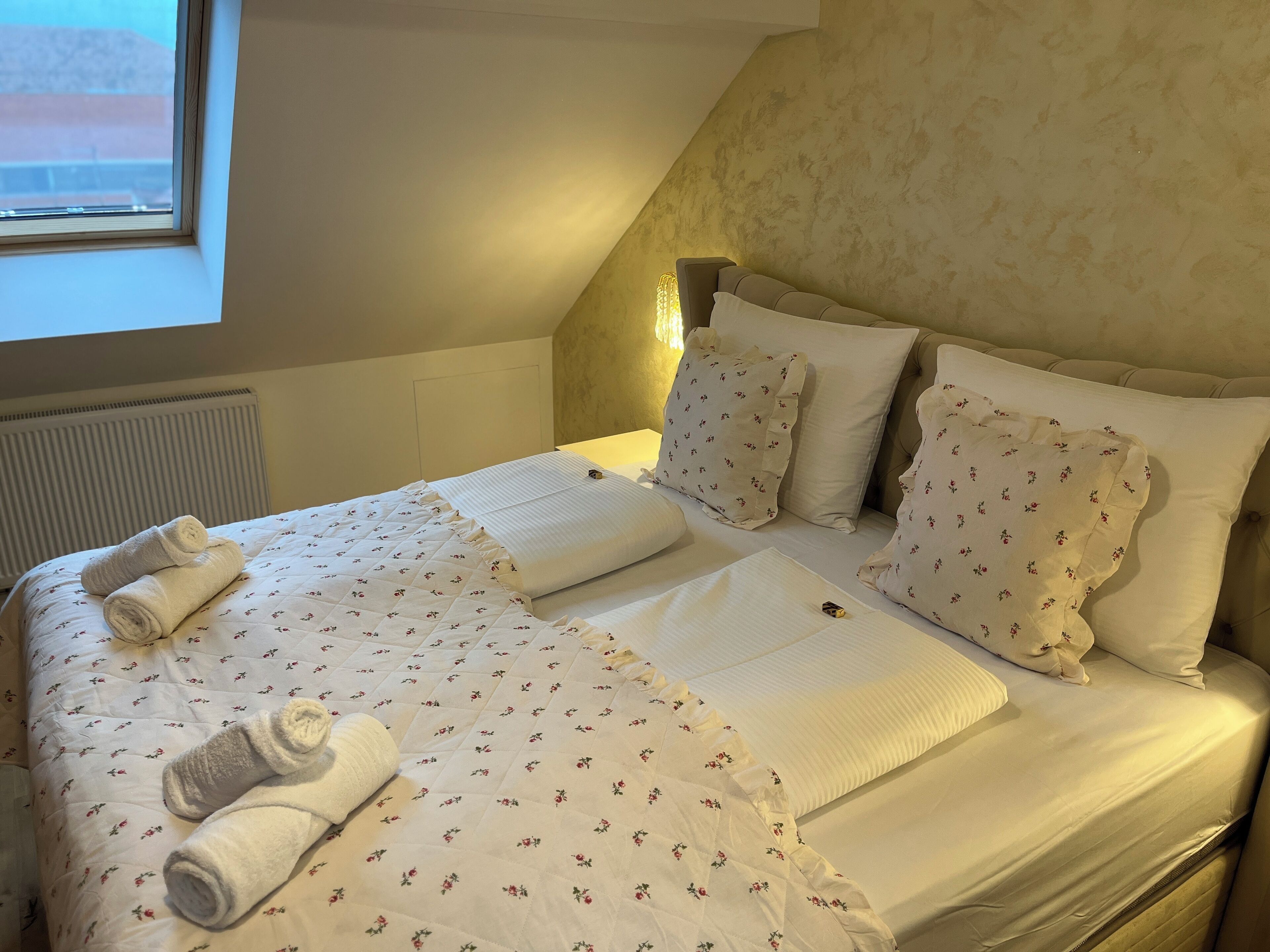 Una cama bien hecha con ropa de cama floral y toallas enrolladas, un radiador y una ventana con vistas.