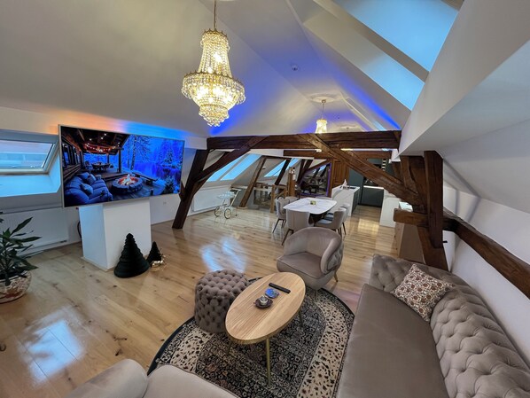 An attic-style living room with a large TV, wooden beams, a chandelier, and a cozy seating area.
