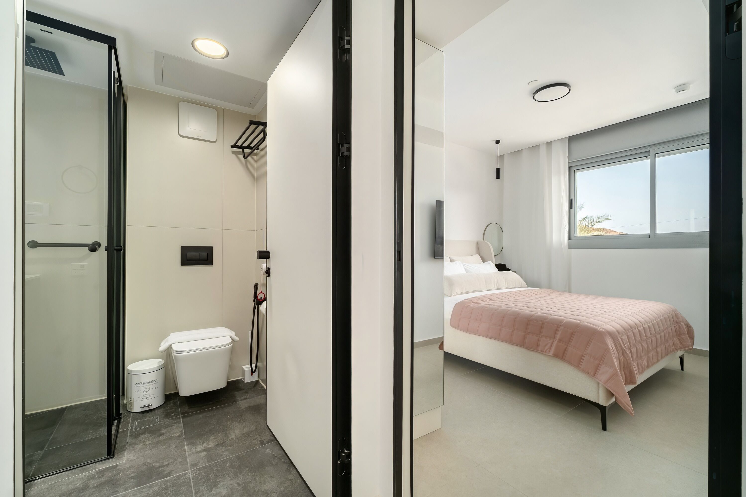 Een moderne badkamer met een douchecabine van glas, een toilet en een bidet. De slaapkamer heeft een bed met een roze deken, een nachtkastje en een raam met uitzicht.