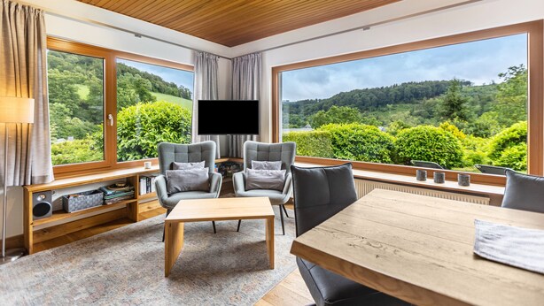 Ein modernes Wohnzimmer mit großem Fernseher, grauen Sesseln, einem Holztisch und Blick auf eine bewaldete Landschaft durch große Fenster.