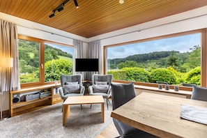 A modern living room with a large TV, grey chairs, a wooden table, and a view of a forested landscape through large windows.