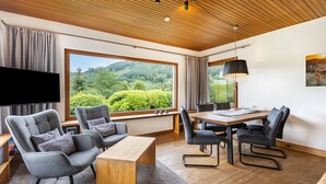 Ein modernes Wohnzimmer mit großem Fenster, Flachbildfernseher, grau gepolsterten Sesseln, einem Holztisch und einem Esstisch mit Stühlen.