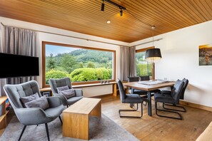 A modern living room with a large window, a flat-screen TV, grey upholstered chairs, a wooden coffee table, and a dining table with chairs.