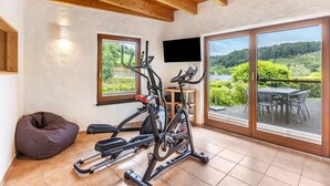 Ein Fitnessraum mit Trainingsgeräten, einem Fernseher und großen Fenstern mit Blick auf Grünflächen.
