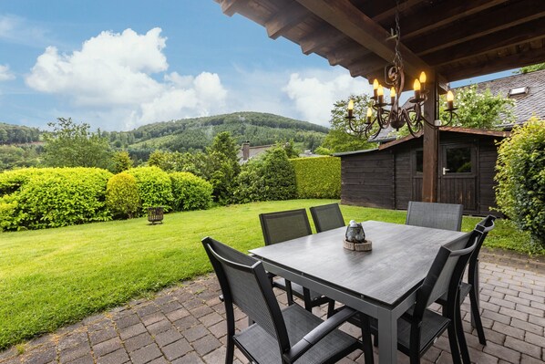 An outdoor patio with a table and chairs, surrounded by a well-maintained lawn and hedges, under a wooden pergola with hanging lights.