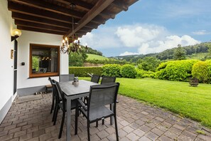 An outdoor patio with a table and chairs, a wooden ceiling, and a view of a green landscape.