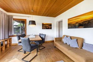 A room with a wooden ceiling, a dining table with chairs, a sofa, and two paintings on the wall.