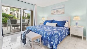 A bedroom with a bed, two bedside tables, a bench, a ceiling fan, and a view of an outdoor patio with chairs and plants. - Ocean Villas Unit 118: Lagoon-Side Luxury with Direct Access (St. Augustine Beach)
