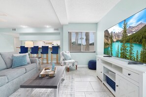 A modern living room with a sofa, coffee table, and a TV displaying a mountain landscape. - Ocean Villas Unit 118: Lagoon-Side Luxury with Direct Access (St. Augustine Beach)