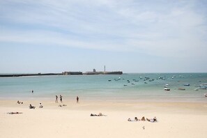 Una playa con agua cristalina, barcos y un faro a lo lejos.