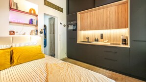 Ein modernes Schlafzimmer mit Bett, Nachttisch und einer integrierten Küchenzeile mit Mikrowelle, Kaffeemaschine und Schrankflächen.