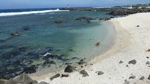 Una playa con agua cristalina, rocas y un faro a lo lejos.