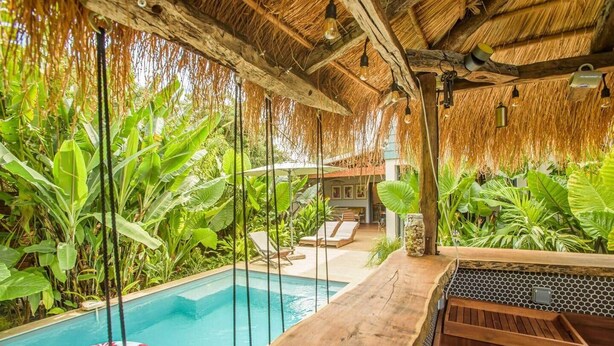 Un área de piscina con terraza de madera, sillas de descanso y techo de paja.