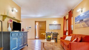 Una sala de estar con un sofá, un televisor sobre un mueble bajo, una mesa de comedor con sillas y un cuadro en la pared.