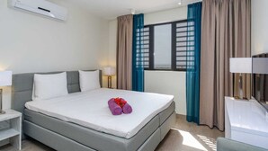 A hotel room with a bed, a desk, a chair, a window with curtains, and an air conditioning unit. - Stylish sea-view apartment with sunset terrace - Coral Estate (Sint Willibrordus)