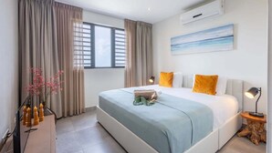 A modern bedroom with a large bed, beige curtains, a window with blinds, a painting on the wall, and a small table with a lamp. - Luxury sea-view apartment with terrace - Coral Estate (Sint Willibrordus)