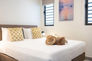 Un lit bien fait, recouvert d’une couette blanche, agrémenté de deux coussins à motifs, d’un chapeau décoratif et d’un petit vase avec des fleurs, dans une chambre d’hôtel.