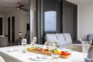 Une table à manger dressée avec des assiettes, des verres et une variété de plats, dont une planche de charcuterie, dans un intérieur moderne agrémenté d’un ventilateur de plafond et de grandes fenêtres.